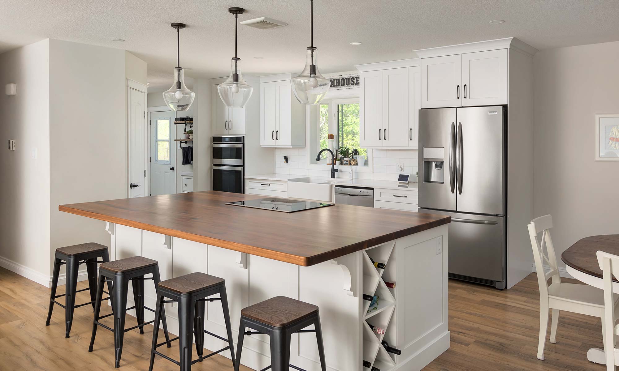 Kitchen with island finished in white featuring a darker wood countertop with inset cooktop and integrated wine rack on the side of the island. Three pendant lights hang above, and four black stools line one side of the island.