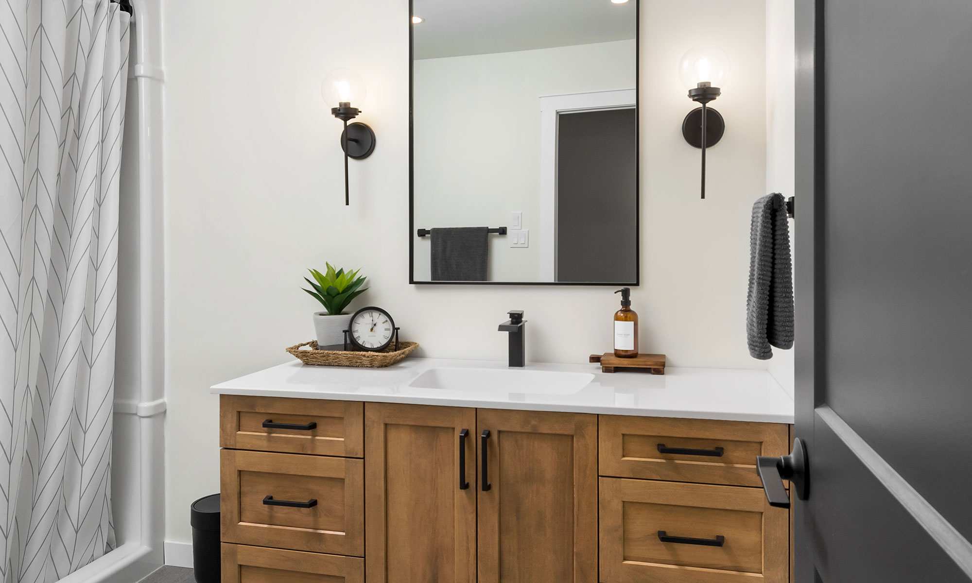 White cultured marble vanity countertop and integrated sink with matte black waterfall faucet. The countertop is installed on a light brown flat-panel vanity with black cabinet pulls. A large mirror is framed by single-light sconce lamps with clear glass globes off to each side. Photo by Illia Sternichuk.