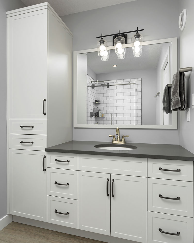 creative-kitchens-mb_portfolio-jeannot_02 A white fibreboard bathroom vanity with two doors and six drawers (3 on each side) is shown with a medium gray countertop and light brass two-handled faucet. A mirrors spans the entire width of the vanity, and a three-lamp light fixture with a black matte finish and clear glass lamp shades hangs above the mirror. To the left of the vanity is a floor to ceiling cabinet with a large door on the top, then two drawers, with a large door below.