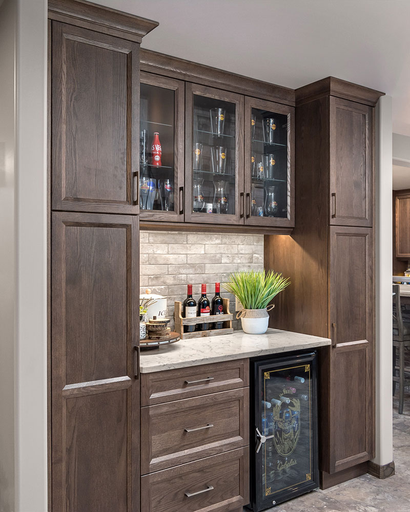 creative-kitchens-mb_portfolio-jeannot_02 A kitchen wine bar with flat-panel cabinets is shown in a medium-brown finish. The centre of the bar features upper and lower cabinets. There are three upper cabinets with glass panel door inserts; below these upper cabinets is a medium gray brick-like backsplash and a matching countertop. Below is a base cabinet with three drawers, with the top drawer narrower than the lower two drawers. A wine fridge sits to the right. Bookending the centre set of cabinets are two floor-to-ceiling match cabinets, each with one upper door that is the height of the upper cabinets, and one lower door which spans the remaining height.