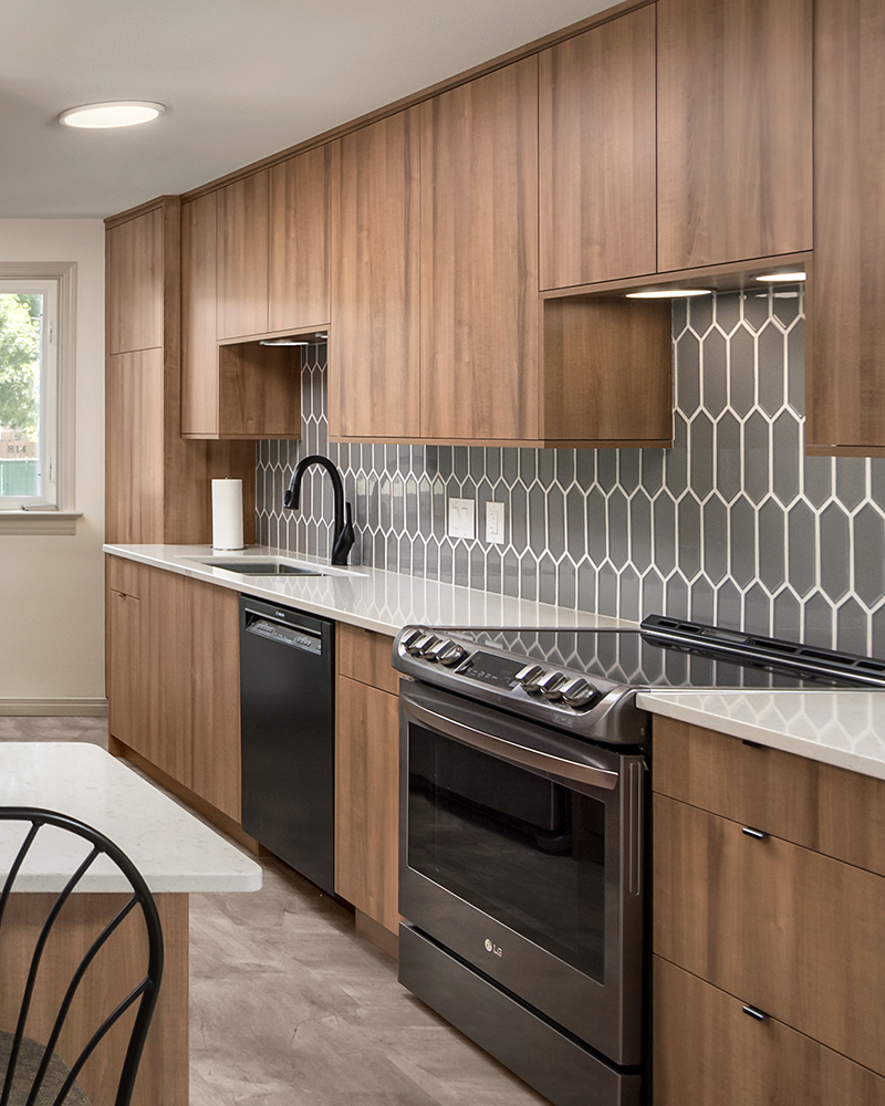 creative-kitchens-mb_portfolio-jeannot_02 A kitchen with melamine cabinets in a medium brown wood pattern. A stainless steel stove is in the foregroud, an undercabinet dishwasher and undermount sink with black-tone faucet is in the background. To the left is an island. All cabinets have white countertops.