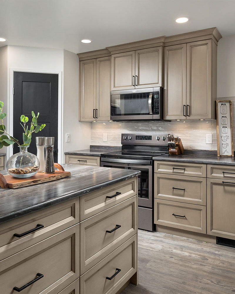 creative-kitchens-mb_portfolio-jeannot_02 A kitchen with beige-coloured ply-panel cabinets with a black and grey stone-pattern countertop. An island is positioned in the lower left corner of the photo, and a row of lower and upper cabinets comprise the back wall. The backsplash is made of high-gloss off-white rectangular tiles. A stainless steel stove has a combination range hood and microwave positioned above it. A black panel door provides access to a pantry at the left of the photo.