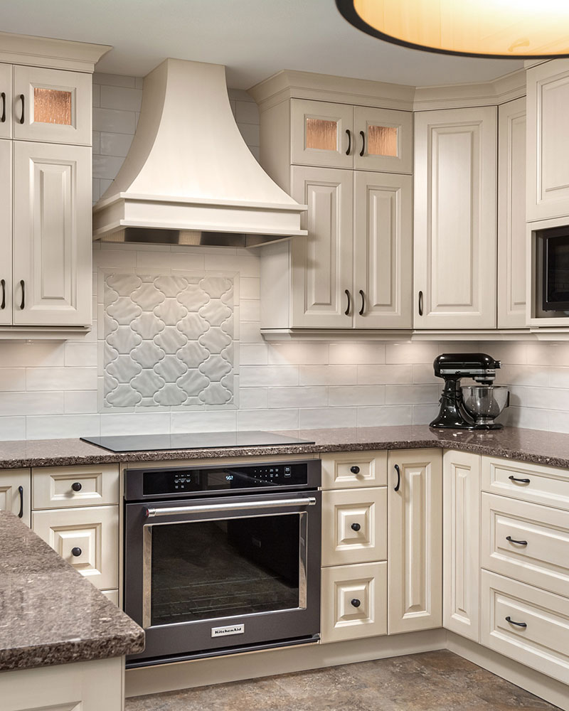 creative-kitchens-mb_portfolio-jeannot_02 A kitchen featuring beige-coloured cabinets with raised panel doors. A stainless steel stove is shown at the centre of the image, with an range hood that matches the cabinet colour.