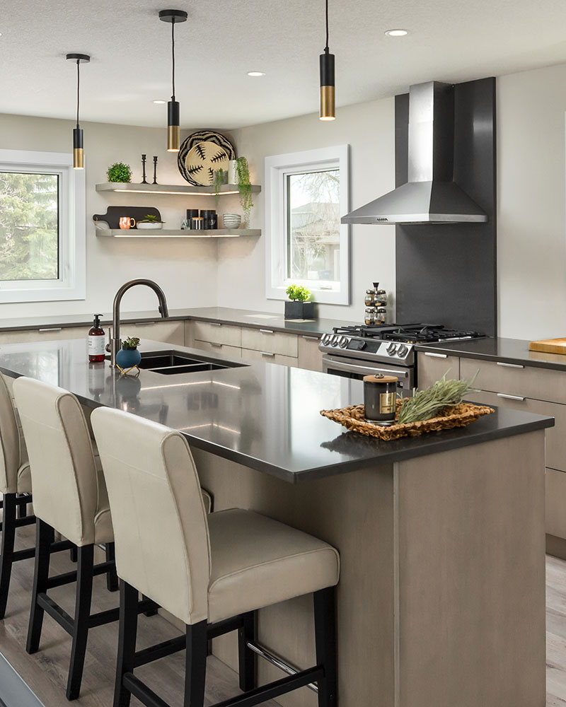 creative-kitchens-mb_portfolio-jeannot_02 A kitchen with slab veneer cabinets is pictured. There are only lower cabinets in a light brown finish, with a medium gray countertop. A set of two L-shaped shelves are in the corner of the kitchen between two white-framed square windows. A stainless steel stove with a gray backsplash and stainless steel range hood is positioned mid-image. Three two-tone (grass and black matte) pendant lights hang from the ceiling.