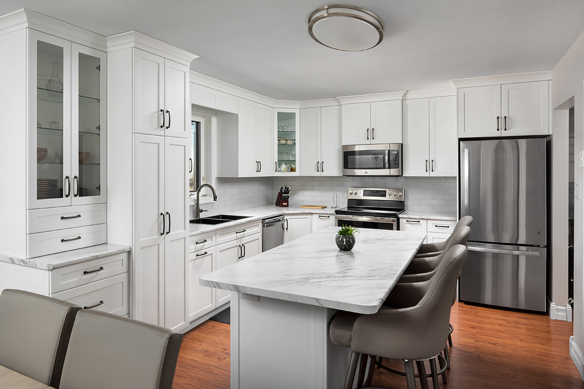 A sample of a kitchen with laminate countertops. The image shows a kitchen with white flat panel cabinets in an L-shaped configuration with an island and stainless steel appliances (stove, refrigerator, microwave and dishwasher). The countertops have a white/gray marble design. Three gray leather upholstered chair stools are arrange along the right side of the island. Along the far left end of the row of cabinets are a floor to ceiling pantry cupboard and a china cabinet with glass door inserts with five drawers below.