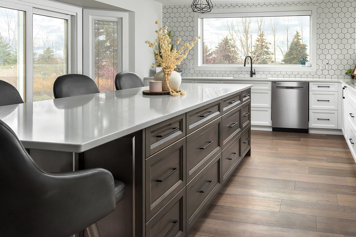 Sample of a quartz countertop. Image shows a kitchen island constructed from dark brown paneled wood with a light gray quartz countertop. The front of the island has a grid of drawers — 4 smaller drawers across the top, with two rows of 4 larger drawers each below. In the background is a counter with a sink against a wall with a large picture window. A backsplash of white hexagonal tiles with black grout extends from counter to ceiling.