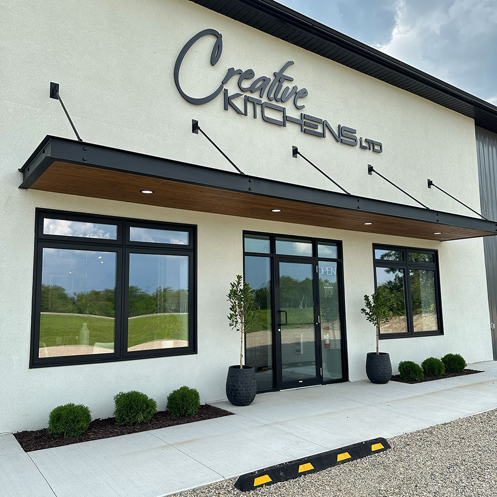 The main entrance to the Creative Kitchens building and showroom at Brandon, Manitoba. The main entrance, at the center of the building, is framed by windows on either side and an awning above. A large sign with the Creative Kitchens logo is affixed to the building above the awning.