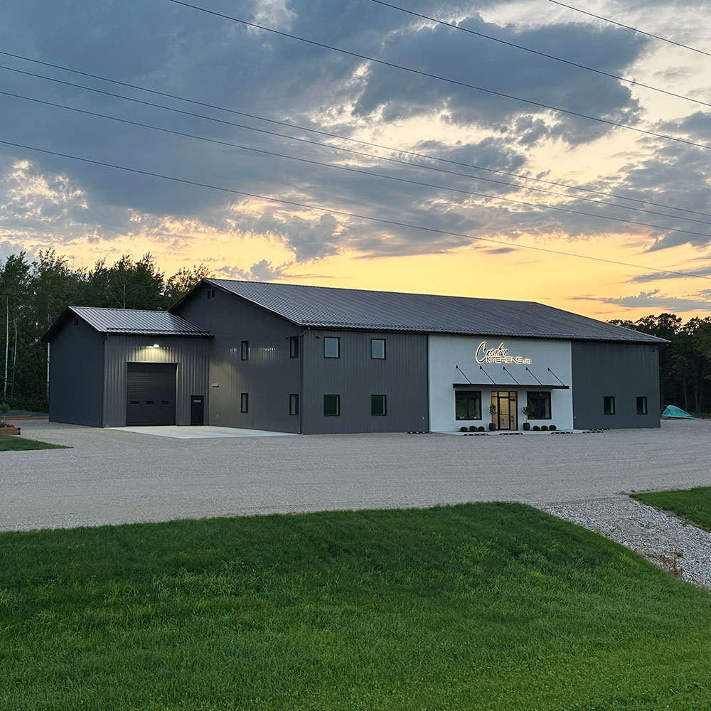 A exterior view of the Creative Kitchens building and showroom at Brandon, Manitoba. The building is black and grey. The main entrance, at the center of the building, is framed by windows on either side and an awning above. A large sign with the Creative Kitchens logo is affixed to the building above the main entrance.